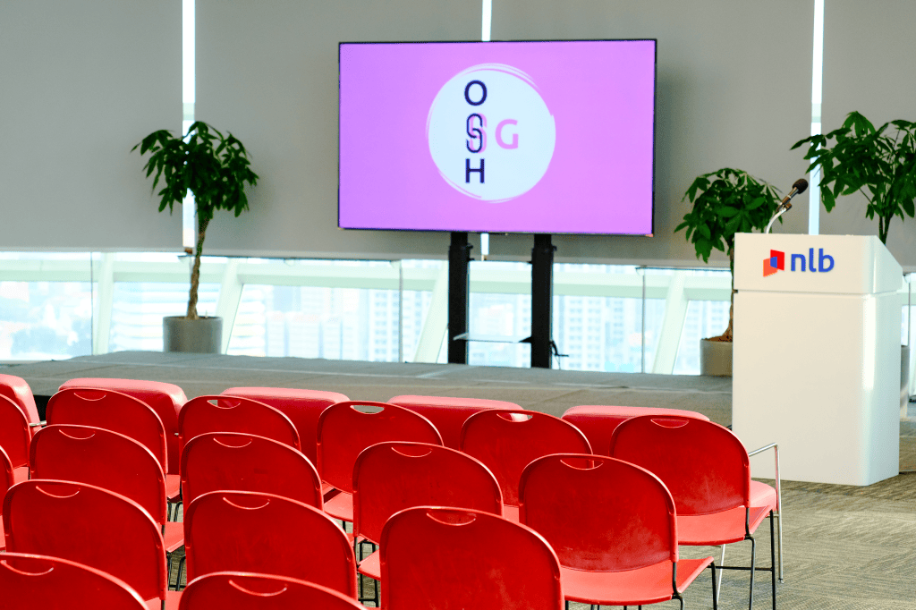 Photo of an event space. In the foreground there are empty red chairs in front of a stage. On the stage there is a pink sign with the OSHSG logo. To the right is a speaking podium with the National Library of Singapore (NLB) logo.